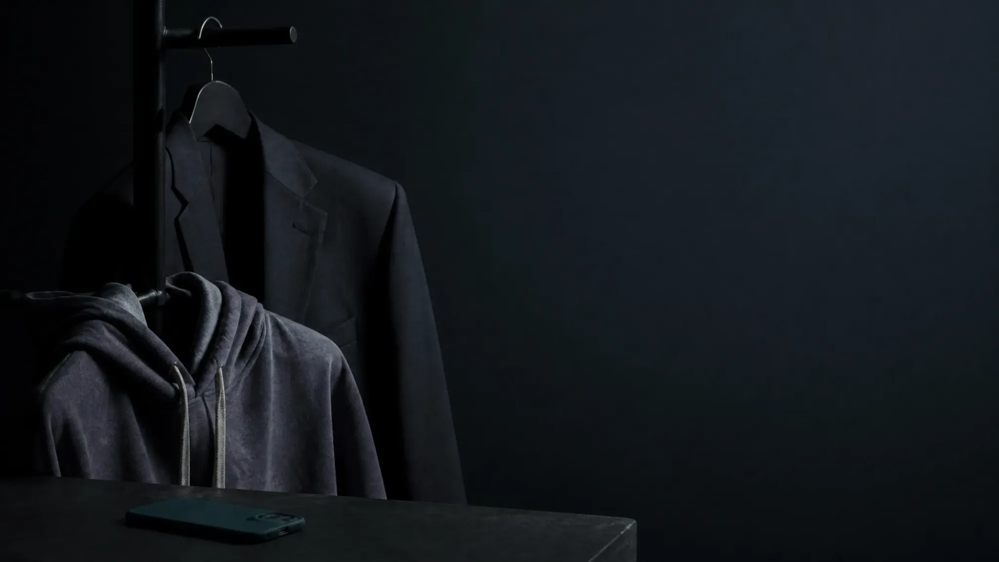 Habitación en penumbra con una chaqueta de traje oscura colgada en un percherero y una sudadera gris con capucha delante, y un móvil con funda verde boca abajo sobre una mesa
