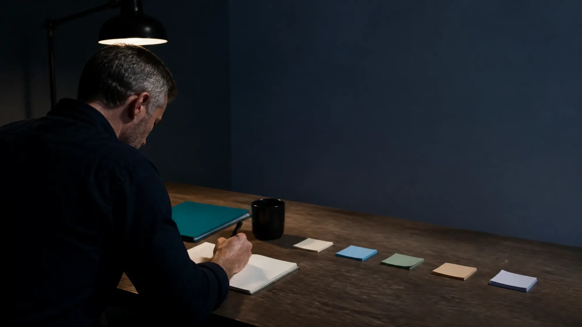 Hombre de pelo canoso, visto de espaldas, escribiendo de noche en un cuaderno bajo la luz de un flexo, con una carpeta verde, una taza negra y cinco tacos de notas de colores alineados sobre la mesa d